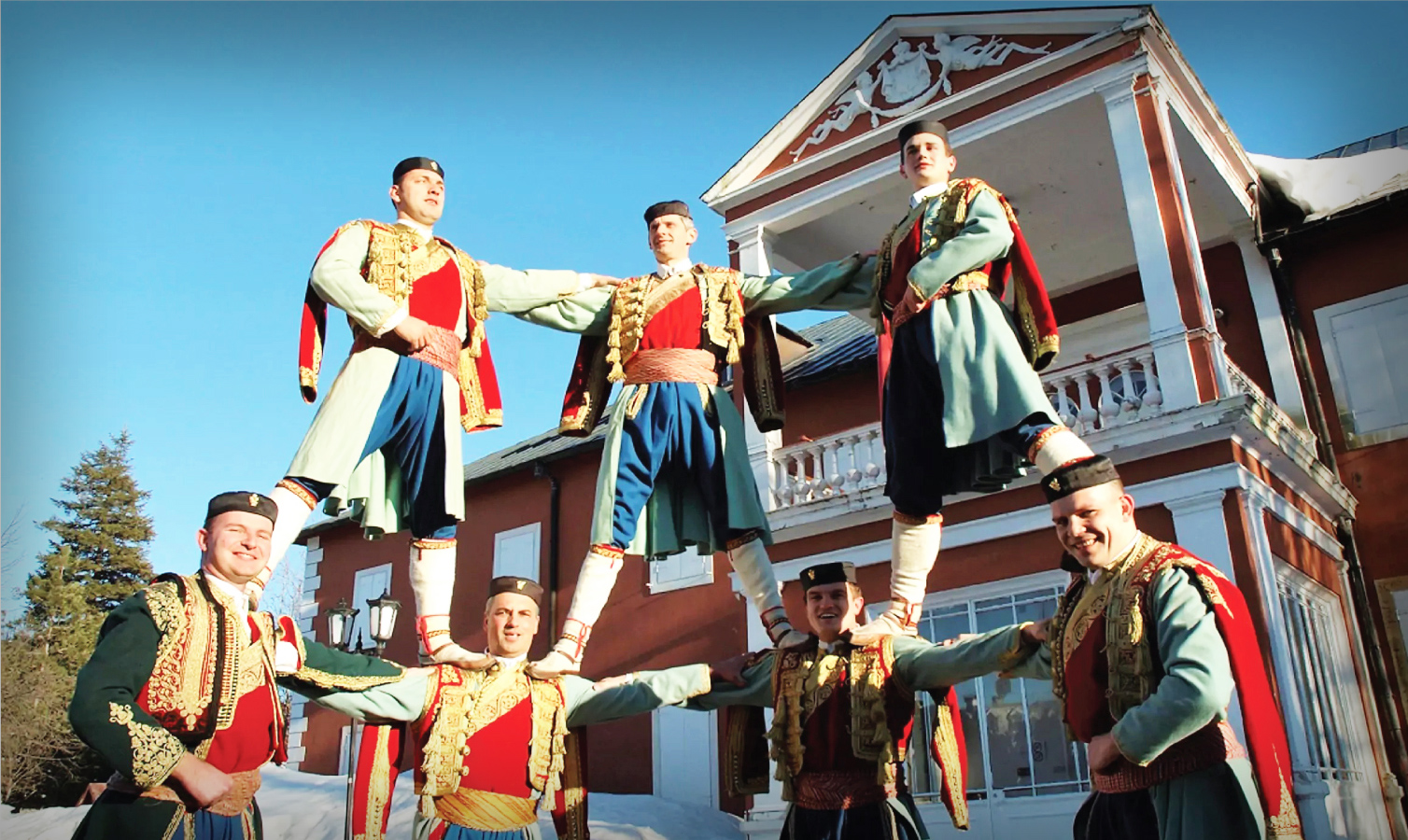 A group of men dressed in traditional Montenegrin folk costumes perform the Oro dance in front of a historic building, standing in a human pyramid formation under a clear blue sky, showcasing cultural heritage and unity.