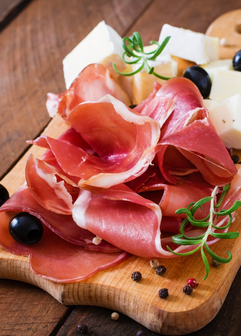 Slices of Njeguši prosciutto arranged on a wooden board, accompanied by pieces of cheese, black olives, peppercorns, and a sprig of rosemary, creating a traditional Montenegrin delicacy presentation.